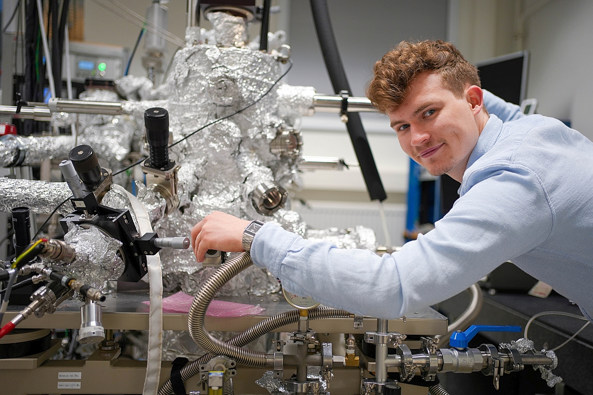 Ein junger mann beugt sich über eine sehr technisch anmutende metallische Apparatur und schaut in die Kamera